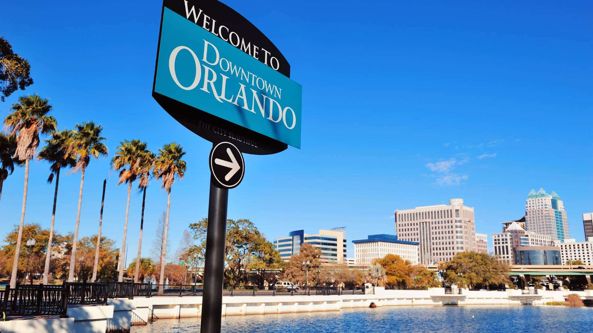 "Welcome to Downtown Orlando" sign with palm trees and city skyline in the background, overlooking Lake Eola.