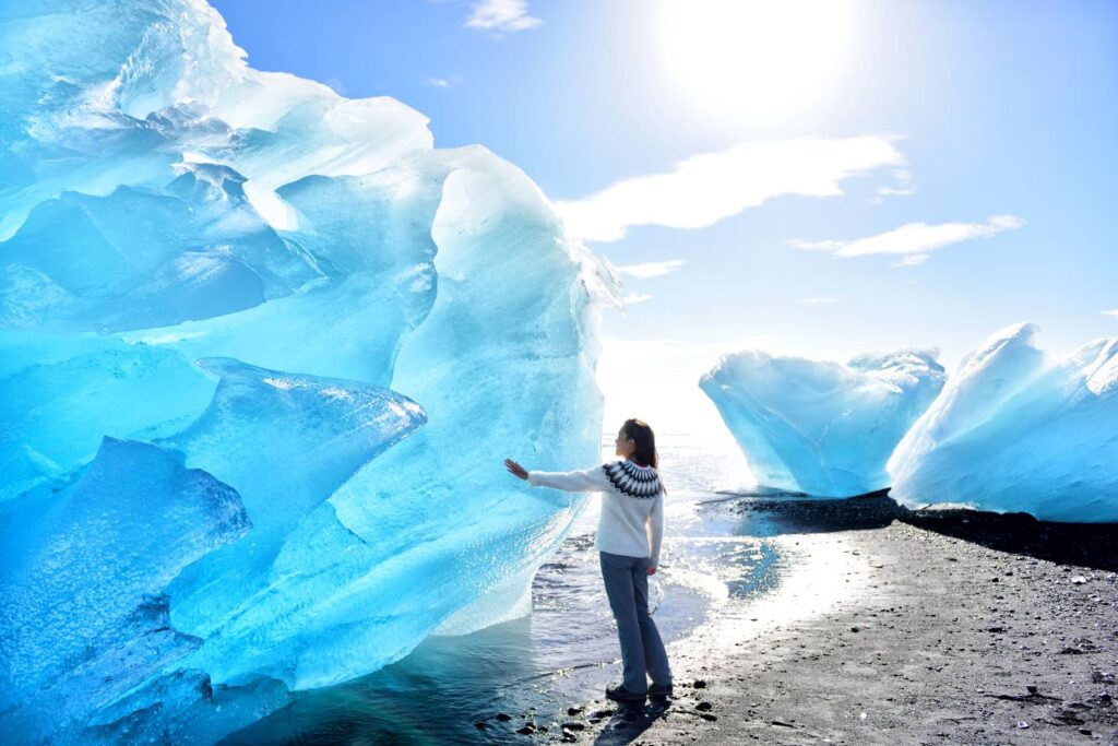 Woman in Nordic sweater touching massive blue iceberg on black sand beach in Iceland, sunlight streaming through translucent ice