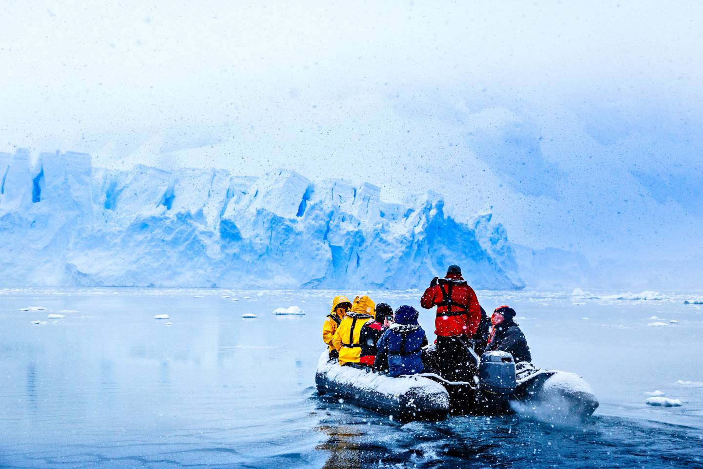 Expedition group in Zodiac boat approaching massive blue glacier wall in Antarctica during snowfall, passengers in colorful winter gear