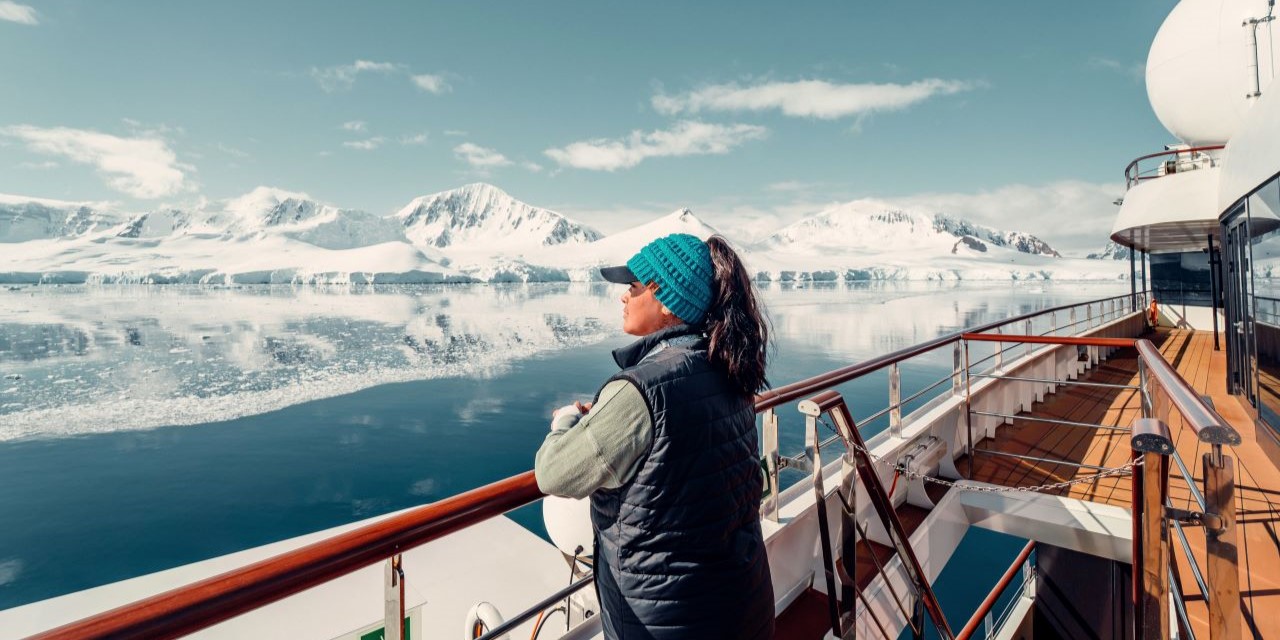 Traveler in blue beanie admiring Antarctic mountain landscape from cruise ship deck, snowy peaks reflecting in calm waters